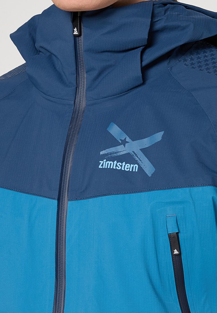 Niebieska i granatowa kurtka przeciwdeszczowa z wysokim kołnierzem, zapięciem na zamek i akcentami z logo. Widoczna teksturowana tkanina oraz detale bocznych kieszeni.