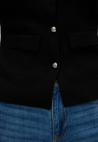 Svart stickad cardigan med silverknappar och två framfickor, tillsammans med blå denimjeans med synlig sömnadsdetalj.