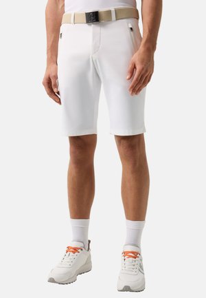 Man draagt witte knielange korte broek met beige riem, witte sokken en witte sneakers met oranje veters tegen een effen achtergrond.
