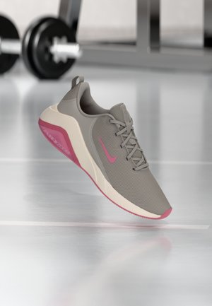 Chaussure de running Nike grise et rose avec lacets, présentée en suspension dans un environnement de salle de sport avec des haltères flous en arrière-plan.