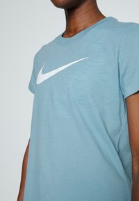 Person bärande en ljusblå kortärmad T-shirt med en stor vit Nike swoosh-logotyp över bröstet mot en enfärgad bakgrund.