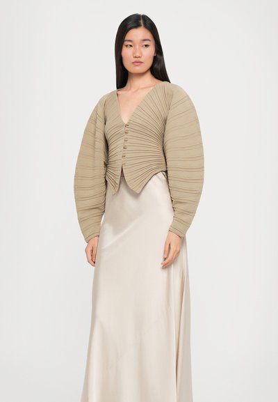 Beige, ribbed cropped cardigan with deep V-neck and large puff sleeves. Features five buttons, worn over a silky, light-colored maxi dress.