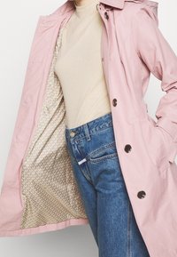 Personne portant un col roulé beige et un jean bleu, montrant la doublure intérieure d'un manteau rose ouvert avec un intérieur beige à motifs.