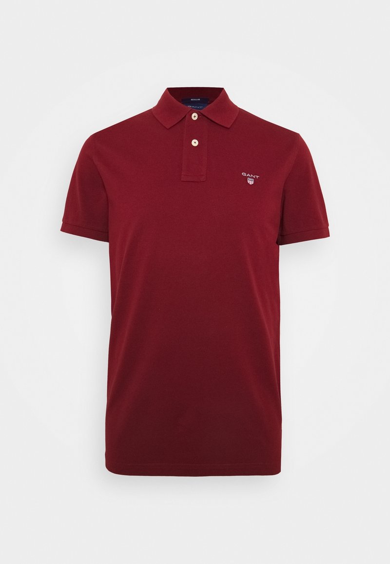Polo rouge foncé à manches courtes avec deux boutons et un petit logo GANT sur la poitrine gauche sur fond blanc uni.