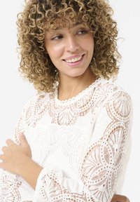 Blusa bianca in pizzo con intricati motivi floreali, scollatura a scalloped e maniche lunghe. Morbida texture con sezioni trasparenti e opache.