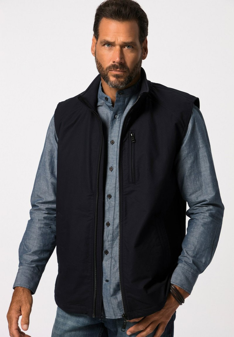 Gilet senza maniche blu navy con zip frontale completa, tasche laterali e tasca sul petto con zip; indossato sopra una camicia azzurro chiaro con bottoni.