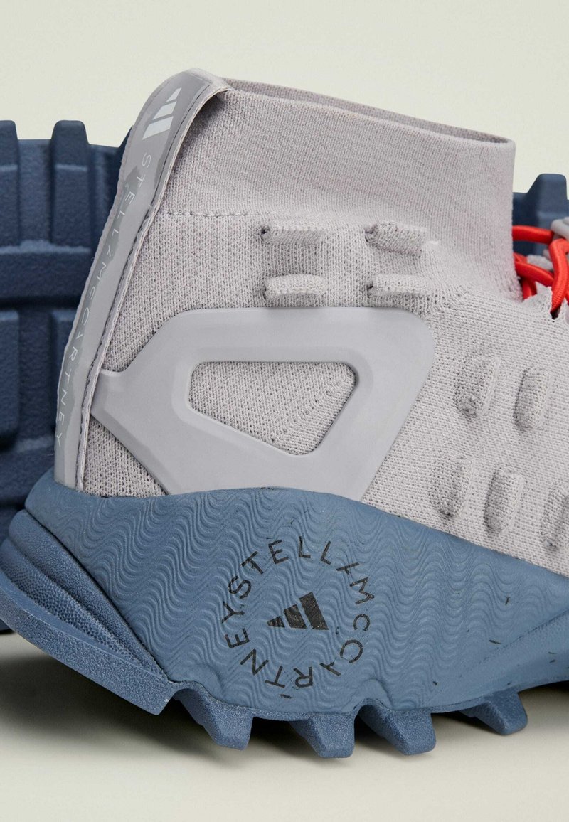 Scarpa sportiva grigio chiaro con tomaia in maglia, dettagli in gomma e suola texturizzata blu. Caratteristiche includono anelli e un logo sul lato.