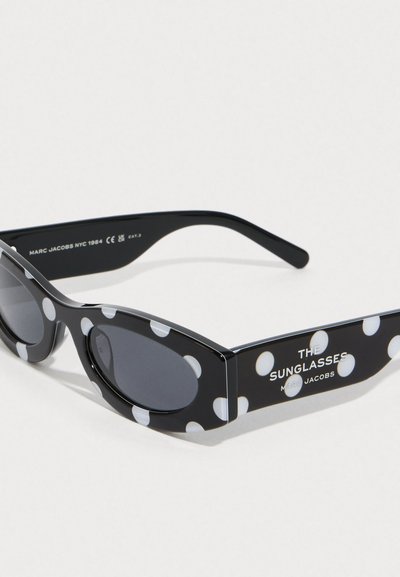 Gafas de sol ovaladas negras con lunares blancos, lentes oscuros y la marca "THE SUNGLASSES MARC JACOBS" en la patilla.