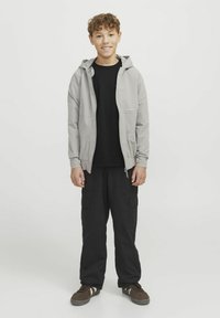 Ljusgrå zip-up jacka med huva, bärs över en svart t-shirt. Svarta cargobyxor och bruna sneakers fulländar outfiten. Enhetlig bakgrund.