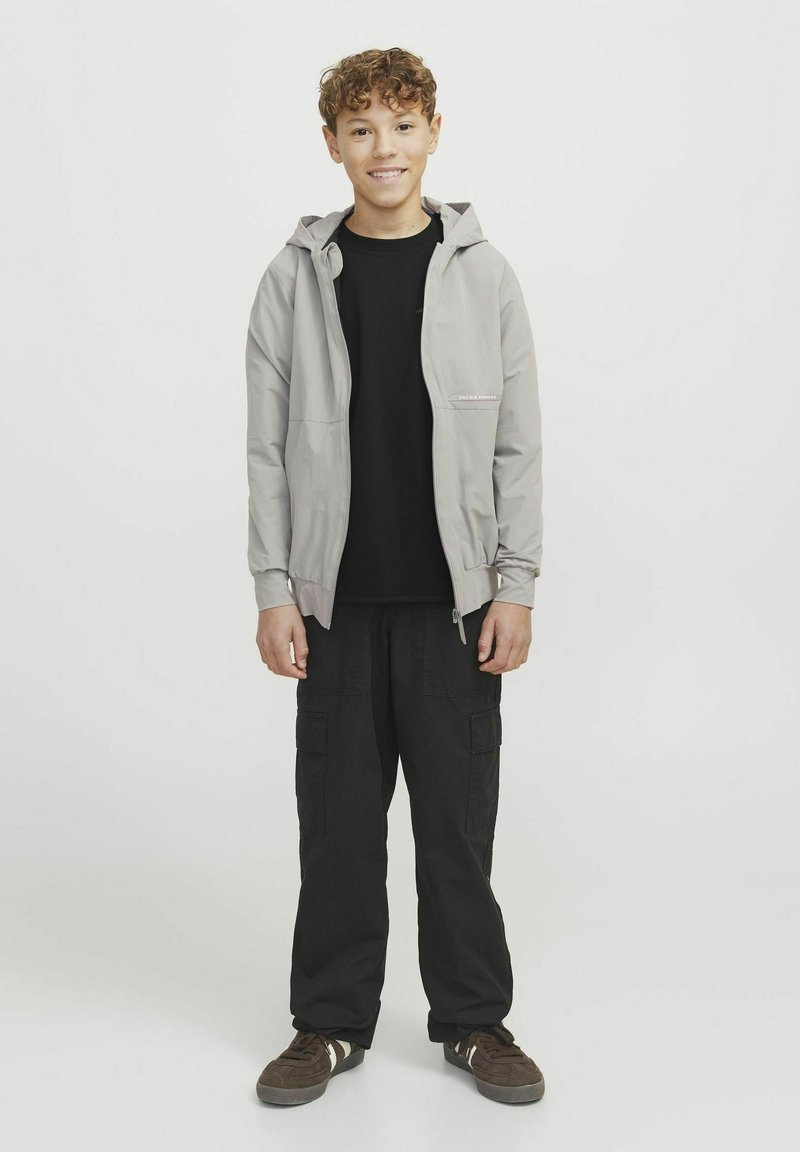 Ljusgrå zip-up jacka med huva, bärs över en svart t-shirt. Svarta cargobyxor och bruna sneakers fulländar outfiten. Enhetlig bakgrund.