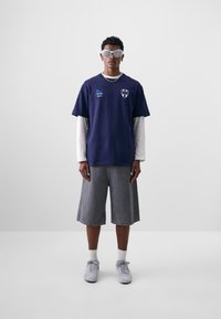 Camiseta de marina con logo, superpuesta a una camiseta de manga larga blanca. Pantalones cortos de mezclilla grises y zapatillas grises, finalizados con gafas de sol de gran tamaño.