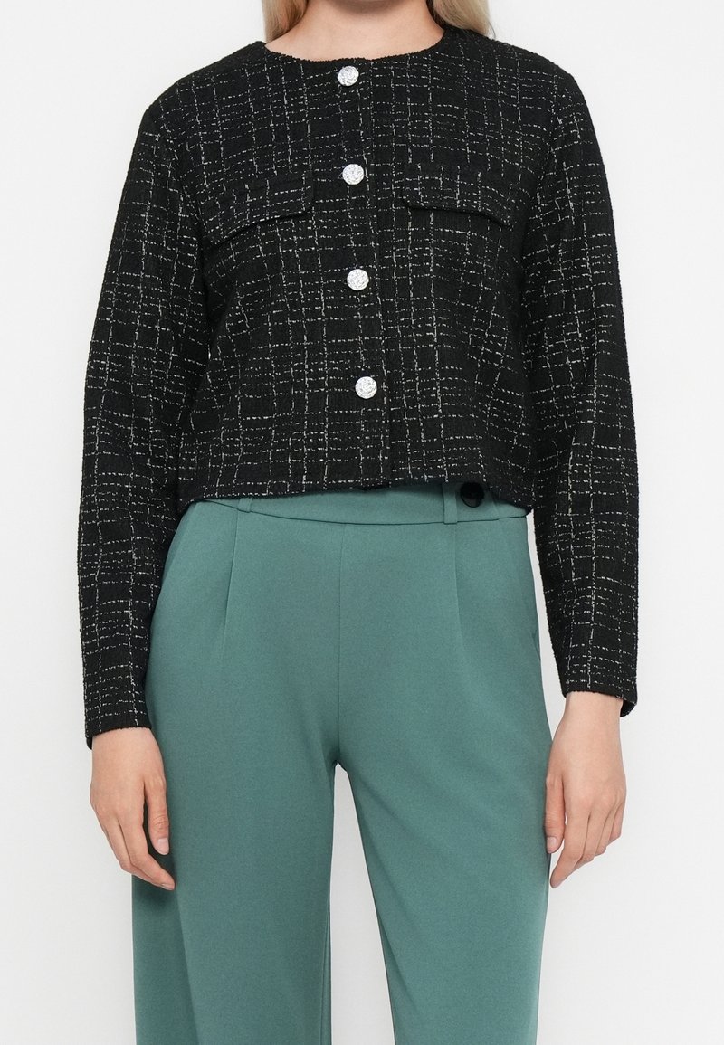 Femme portant une veste courte texturée noire à boutons argentés, associée à un pantalon large taille haute couleur sarcelle.