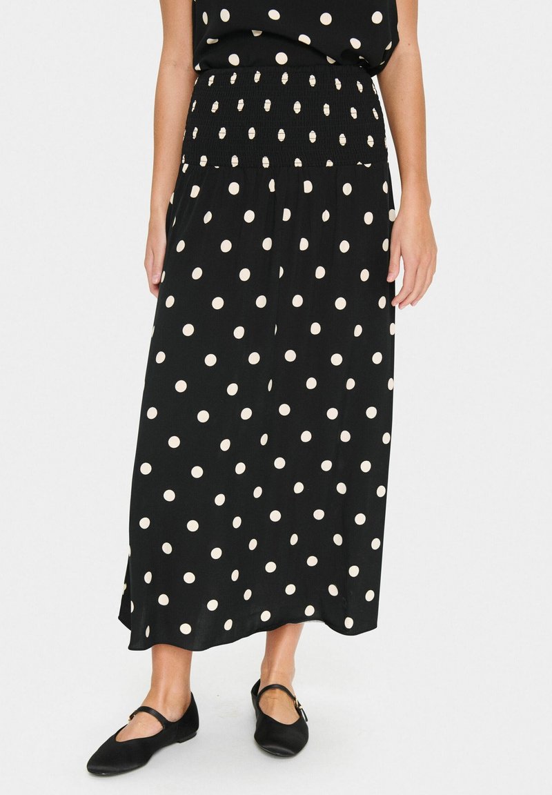 Femme portant une jupe midi noire à pois blancs, associée à des ballerines Mary Jane noires, debout devant un fond blanc.