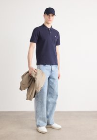 Marinblå pikétröja med krage, ljusblå baggy jeans och vita sneakers. Håller en beige jacka och har en marinblå keps på sig. Slät tygkvalitet.