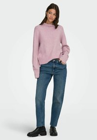 Kvinne iført en rosa genser med perledetaljer på kragen og mansjettene, blå jeans og svarte kraftige støvler, står med den ene hånden i lommen.