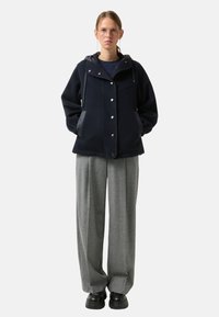 Granatowa kurtka z mieszanki wełny z kapturem, zatrzaskami i dwoma bocznymi kieszeniami; zestawiona z jasnoszarymi plisowanymi spodniami i czarnymi butami na platformie.