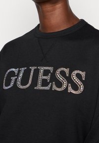 Sweat-shirt noir à col rond avec le lettrage "GUESS" composé de petits strass réfléchissants sur la poitrine, porté par une personne à la peau foncée.