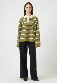 Kvinne iført en olivengrønn og hvit stripete oversized genser med krage og knapper, svarte vide bukser og mørke sko.