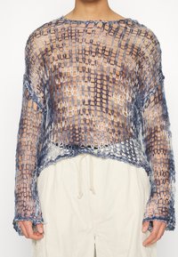 Personne portant un pull en maille bleue et blanche, ample et ouverte, sur peau nue, assorti d’un pantalon clair à cordon.