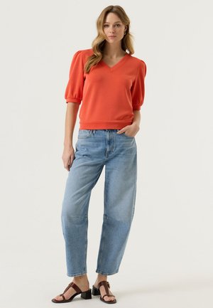 Donna con maglia arancione con maniche a sbuffo a scollo a V, jeans a vita alta azzurri chiaro e sandali con tacco marroni, in piedi con una mano in tasca.