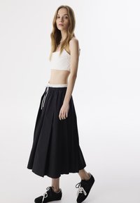 Witte cropped top van textuurstof, gecombineerd met een zwarte geplooide maxi rok met een elastische tailleband en trekkoord. Draagt bij zwarte sneakers.