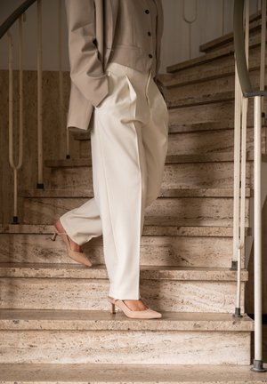 Blazer beige sopra una blusa grigia chiara, abbinato a pantaloni beige larghi e décolleté nudi a punta. Lo sfondo presenta scale in pietra.