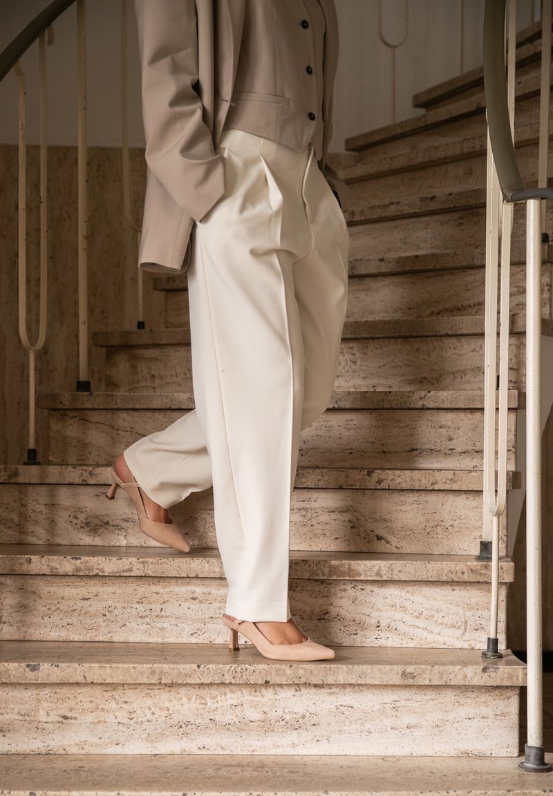 Beige blazer over een lichtgrijze blouse, gecombineerd met losse, crèmekleurige broeken en puntige naaktkleurige hakken. De achtergrond toont stenen trappen.
