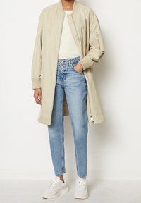 Beige lichte bomberjack met geribbelde manchetten, gedragen over een witte geribbelde top. Gecombineerd met lichtblauwe rechttoe-rechtaan jeans en witte sneakers.