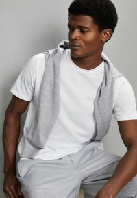 Mand sidder op ad grå baggrund iført hvid t-shirt, lysegrå bukser og grå sweater hængende over skuldrene, ser til siden.