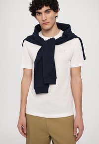 Biela tričko s svetlomodrým logom, doplnené námorníckou mikinou zviazanou okolo ramien. Model má na sebe béžové nohavice.
