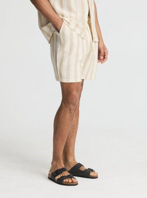 Uomo che indossa pantaloncini a maglia beige a righe e top abbinato, in piedi con una mano in tasca e sandali neri con fibbia.