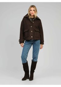 Femme portant une veste boutonnée marron foncé, un jean bleu et des bottes noires jusqu'aux genoux, se tenant devant un fond gris clair uni.