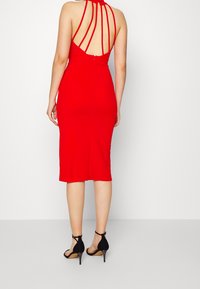 Robe rouge ajustée avec un col halter et un dos nu, comportant de multiples bretelles à l'arrière. Portée avec des sandales à talons hauts noires. Tissu lisse.