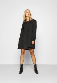 Ξανθιά γυναίκα στέκεται με αυτοπεποίθηση φορώντας μία μακριά μαύρη σακάκι πάνω από ένα μαύρο φόρεμα και μαύρες μπότες μέχρι τον αστράγαλο, μπροστά σε ένα απλό ανοιχτό φόντο.