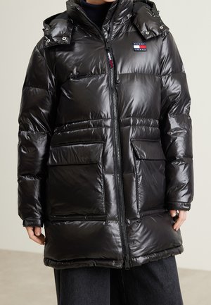 Personne portant une longue doudoune noire brillante avec de grandes poches avant et un logo Tommy Jeans visible sur la poitrine.