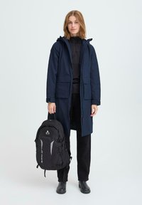 Mornarska dolga jakna s kapuco in več žepi, oblečena preko črne majice in temnih hlač. Črna nahrbtnik z odsevnimi detajli.