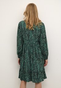 Rochie verde cu model, cu mâneci lungi, talie strânsă și tiv în straturi. Prezintă un amestec de desene abstracte în nuanțe de verde deschis și închis.