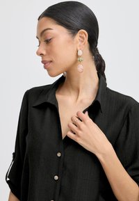 Camicia nera con bottoni e maniche arrotolate. Orecchini in oro e gemme dai colori pastello. La texture liscia e il design aderente mettono in risalto le caratteristiche.