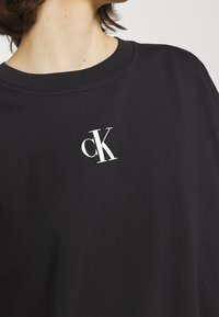 Primer plano de una persona vistiendo una camiseta negra de cuello redondo con un pequeño logo blanco "cK" en el pecho.