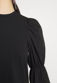 Top nero con maniche lunghe a palloncino e polsini arricciati elasticizzati, indossato con capelli lunghi e scuri e un orecchino pendente colorato con strass.
