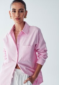 Donna che indossa una camicia rosa a bottoni larga con maniche arrotolate e pantaloni bianchi con cordoncino, in piedi con una mano in tasca.
