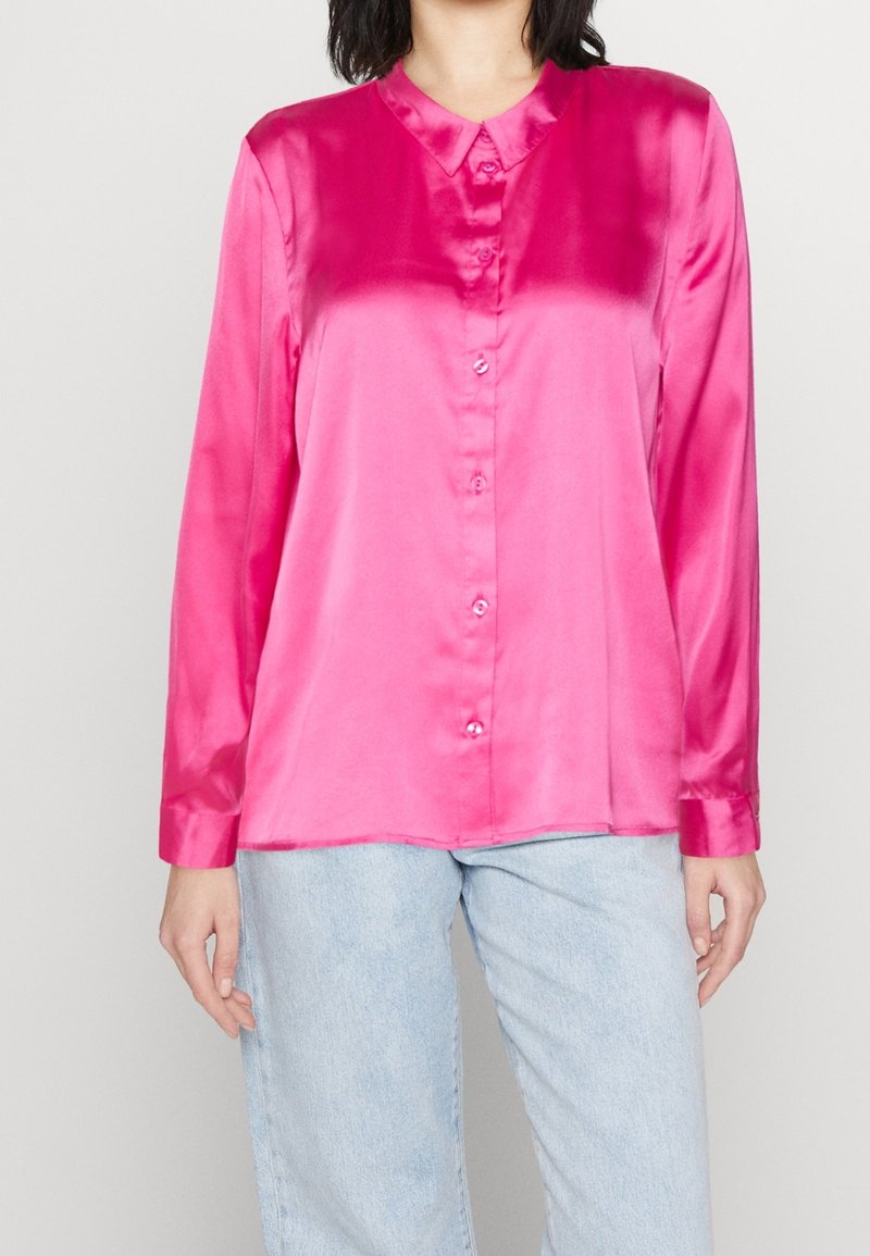 Femme portant une chemise en satin rose vif à manches longues boutonnée, associée à un jean en denim bleu clair, debout devant un fond uni.