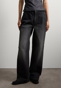 Pantalones de mezclilla negros de tiro alto y pierna ancha con una textura suave y un diseño clásico de cinco bolsillos, combinados con zapatos negros de estilo mocasín.