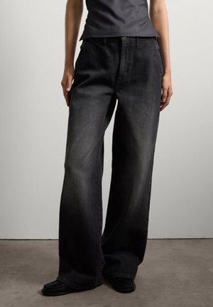 Pantaloni in denim neri a vita alta e gamba larga, con una texture liscia e un classico design a cinque tasche, abbinati a scarpe nere stile mocassino.