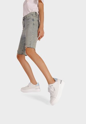 Shorts in denim azzurro con bordi sfilacciati, abbinati a sneakers bianche con suola lucida bianca e un accento azzurro chiaro nel tallone.
