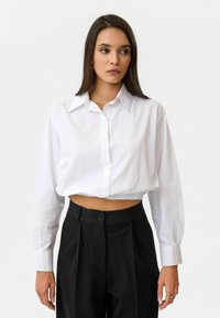 Geknipte witte overhemeld met lange mouwen en een kraag, gedragen met hooggeprofileerde zwarte broek met een voorknop en plooien.