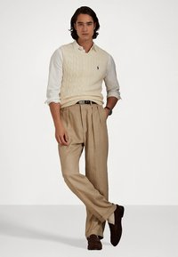 Εκρού γιλέκο με πλεκτό σχέδιο πάνω από λευκό πουκάμισο, συνδυασμένο με μπεζ παντελόνι με πιέτες και καφέ παπούτσια από σουέντ. Απλό ρολόι στον καρπό.