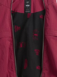 Interiorul unei jachete cărămizii cu căptușeală neagră, prezentând sigle imprimate roșii: "GHP", "Stranger Things" și "Hellfire Club". Se vede un fermoar argintiu.