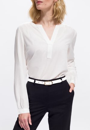 Femme portant un chemisier blanc à manches longues et col en V, rentré dans un pantalon noir avec une ceinture blanche, une main dans la poche, sur un fond uni.