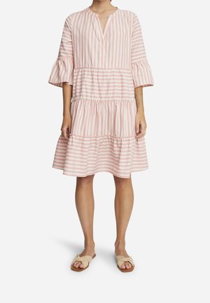 Kobieta w różowo-białej sukience w paski do kolan z warstwową spódnicą i falbaniastymi rękawami do łokcia, do tego beżowe sandały wsuwane.